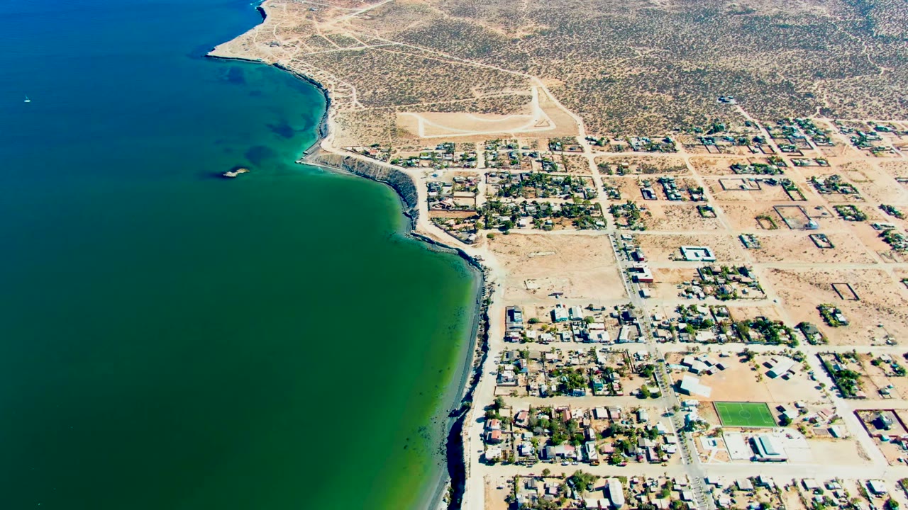 Mid-clip aerial frame over San Juanico showing the bay on the left, sandy shoreline and darker rocks at the waterline, a compact town grid, a green sports field, desert tracks, and no visible people.