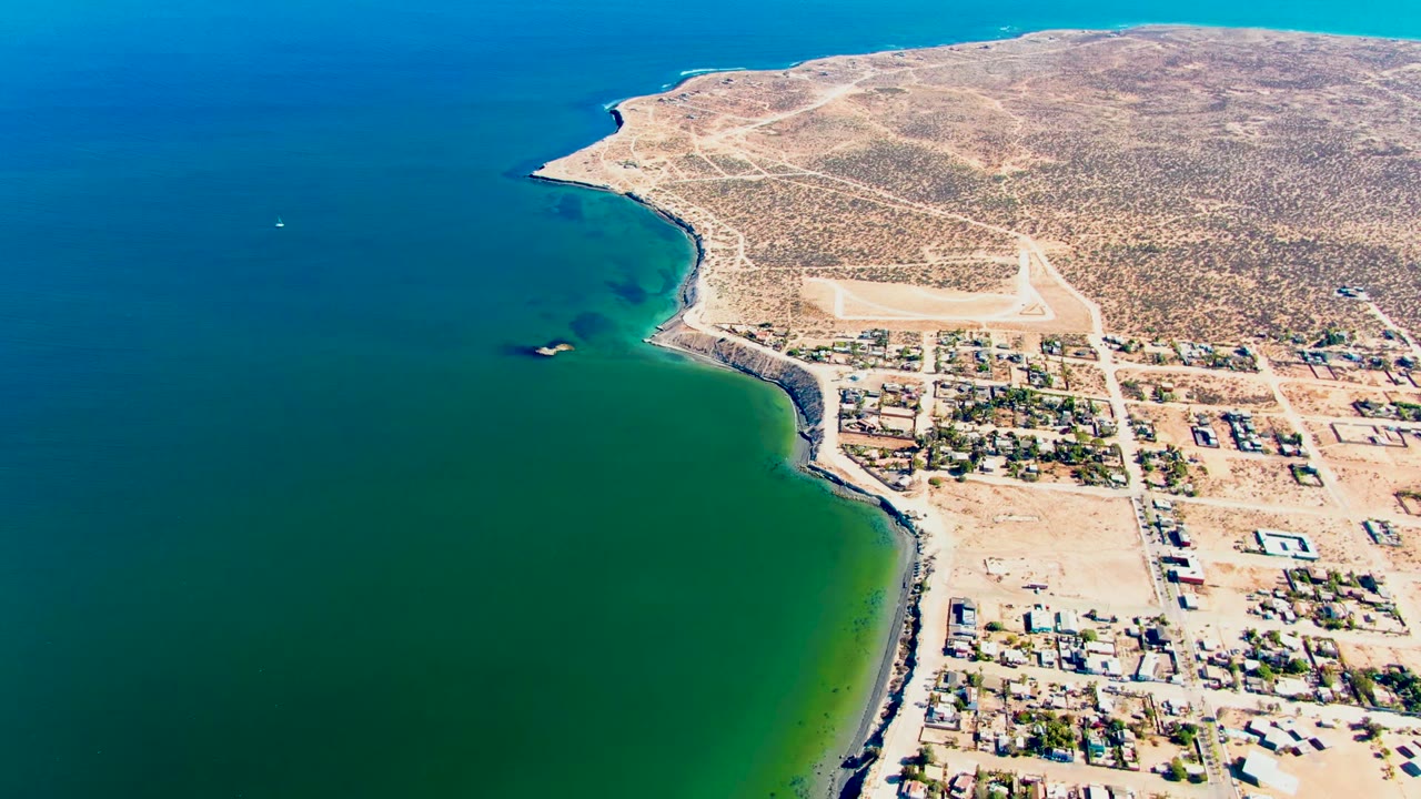 High aerial view over San Juanico, Baja California Sur, with green-blue water on the left, sandy town blocks on the right, desert hillside beyond, and no visible people.