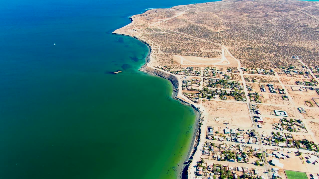 Aerial view over San Juanico showing the boat farther across the water, the sandy road grid, scattered homes, a green field at lower right, and no visible people.