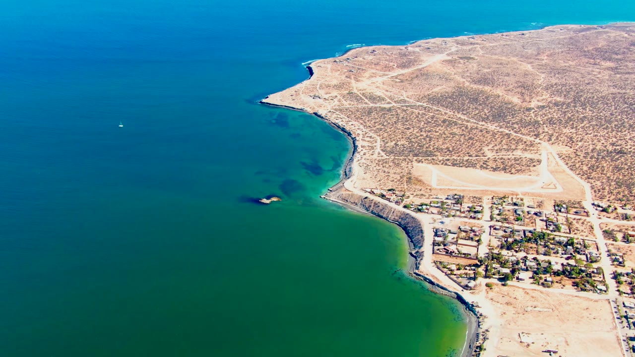 Opening aerial frame over San Juanico, Baja California Sur, with blue-green water on the left, a rocky desert shoreline, roads, and small coastal buildings, with no visible people.