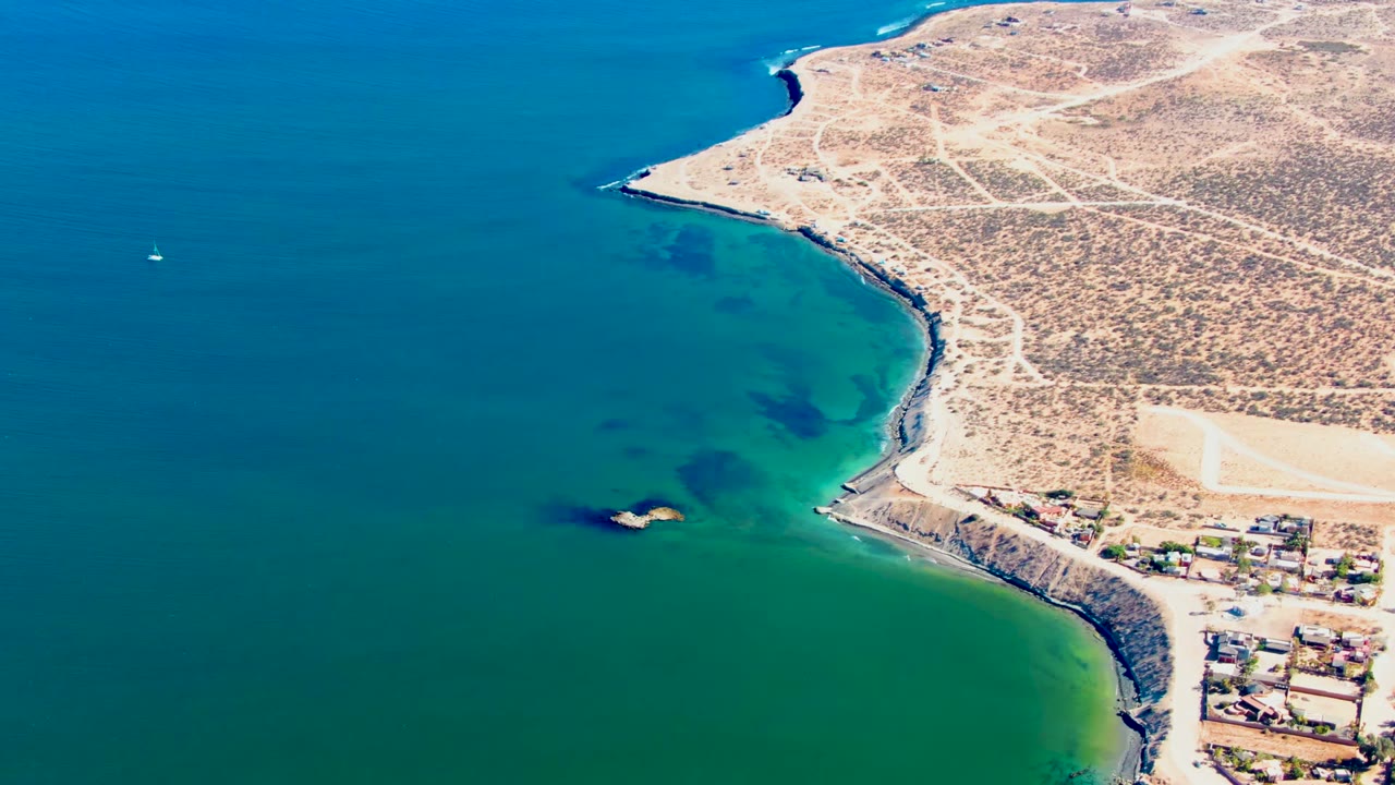 Aerial frame over San Juanico, Baja California Sur showing blue-green water, a rocky shoreline, dry tan land with dirt roads, a small settlement at lower right, and no visible people.