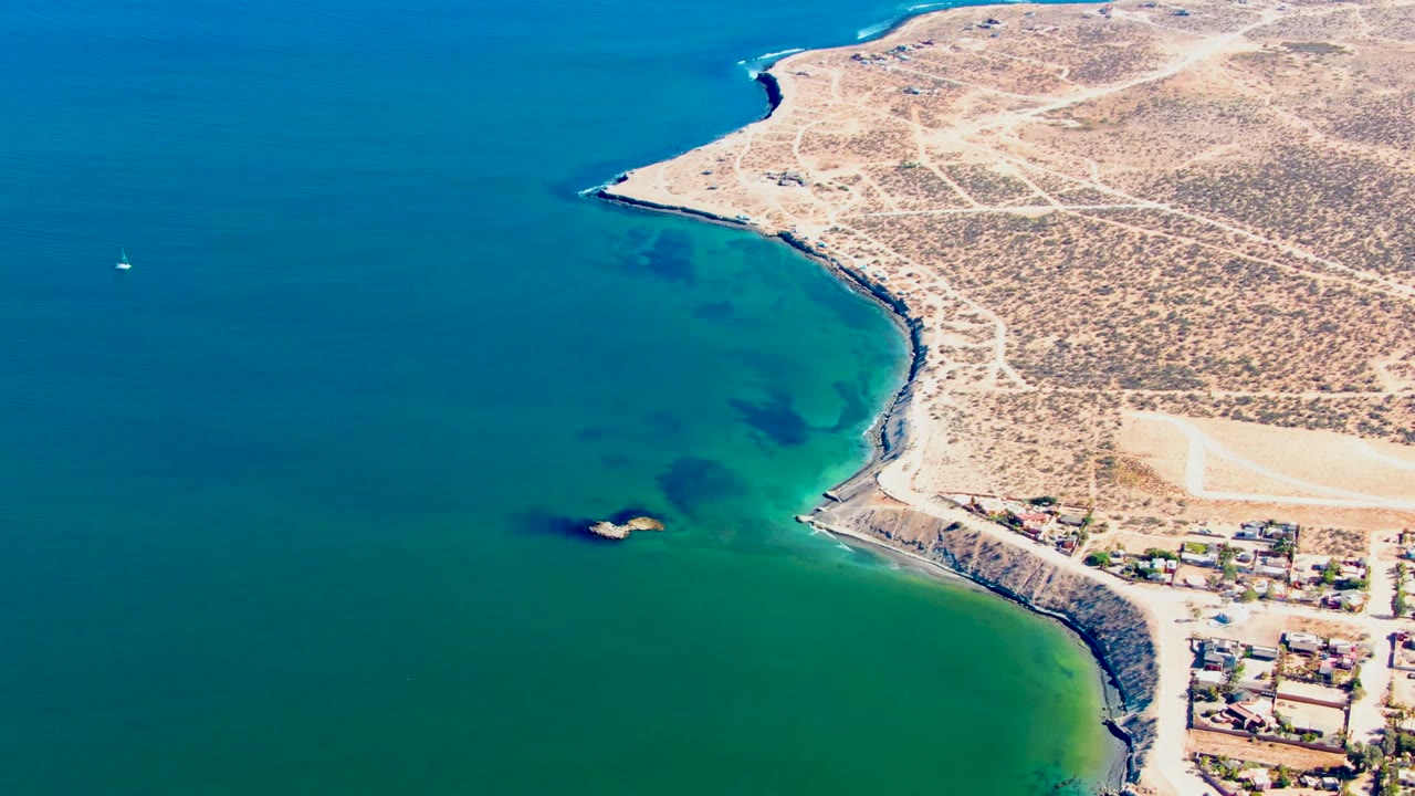 San Juanico coastline seen from above, with green water in the bay, a tan headland, pale dirt roads crossing arid ground, buildings along the lower right edge, and no visible people.