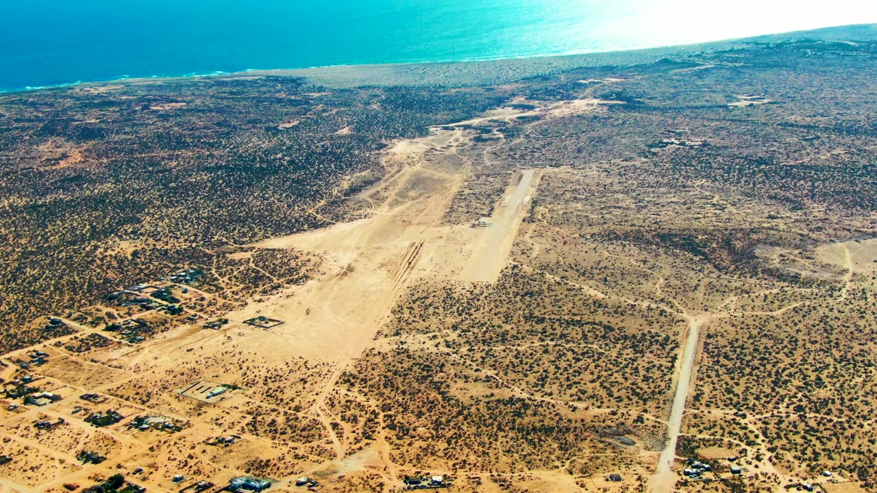High aerial frame over San Juanico, Baja California Sur, showing a sandy runway through scrub desert, scattered small buildings near the lower left, blue ocean on the horizon, and no visible people.