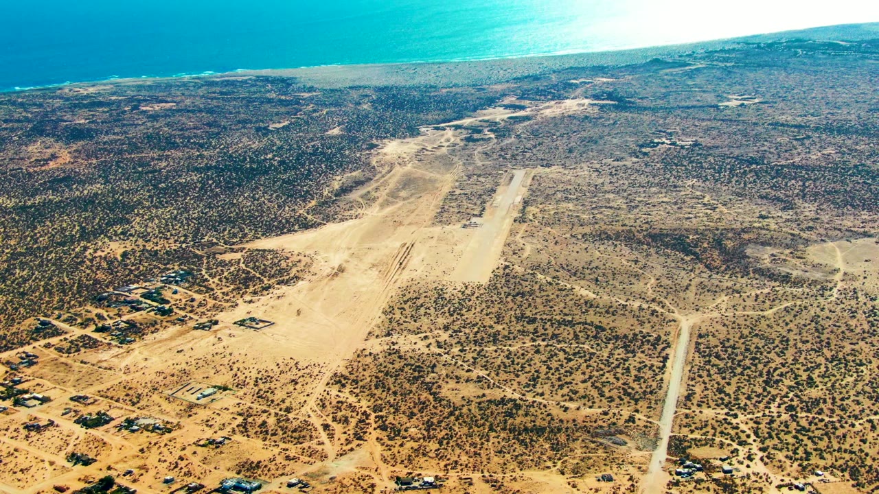 Aerial view in San Juanico, Baja California Sur, with the runway centered slightly right, branching dirt tracks across tan scrubland, coastal water across the top edge, and no visible people.