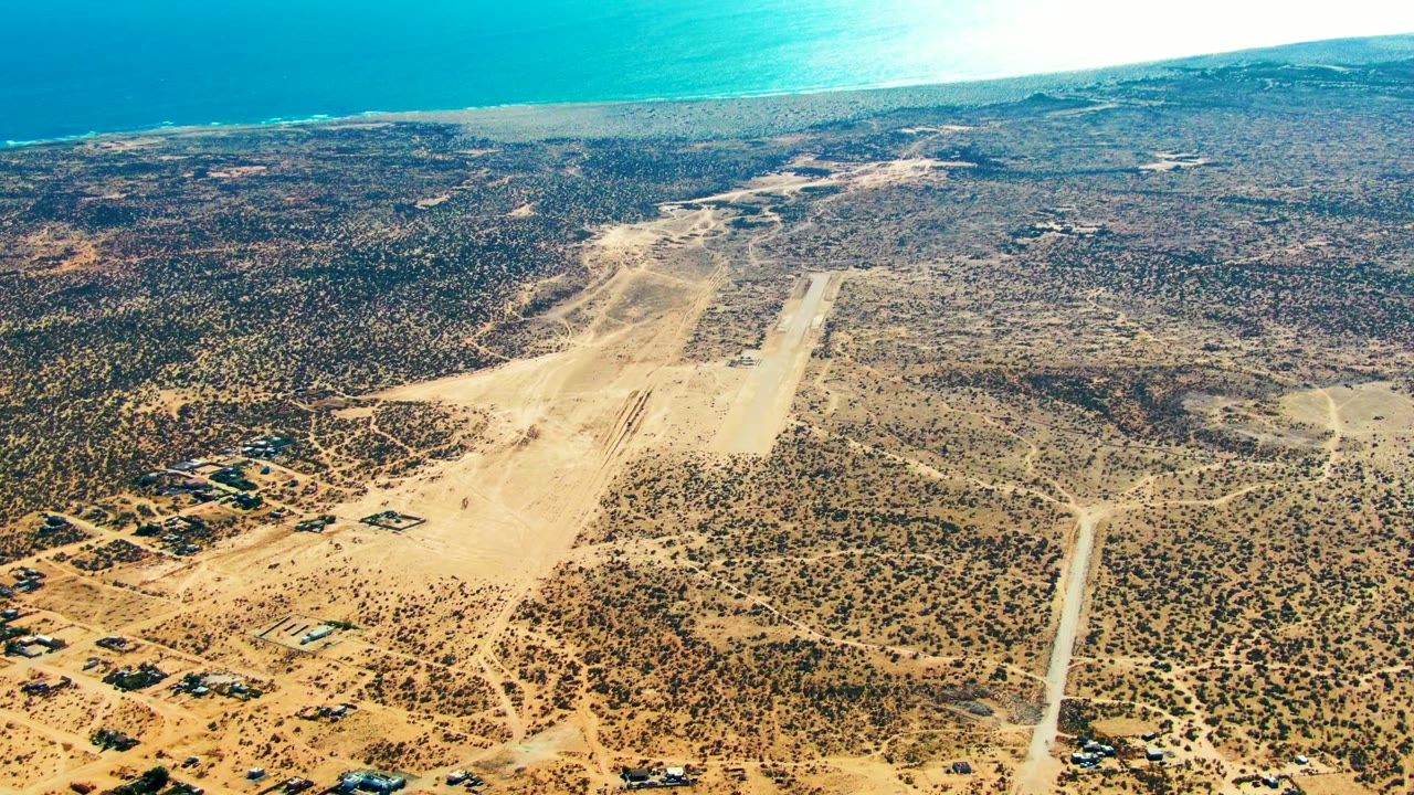 Wide aerial frame over San Juanico, Baja California Sur, showing the pale desert runway, darker scrub patches, scattered low buildings at the lower left, turquoise ocean in the distance, and no visible people.