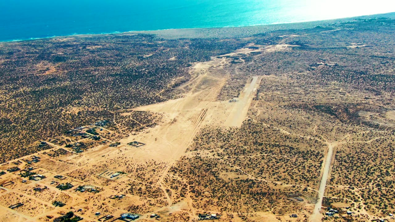 Aerial frame over San Juanico, Baja California Sur, showing a sandy runway, dirt roads, sparse desert vegetation, scattered buildings, blue ocean in the distance, and no visible people.