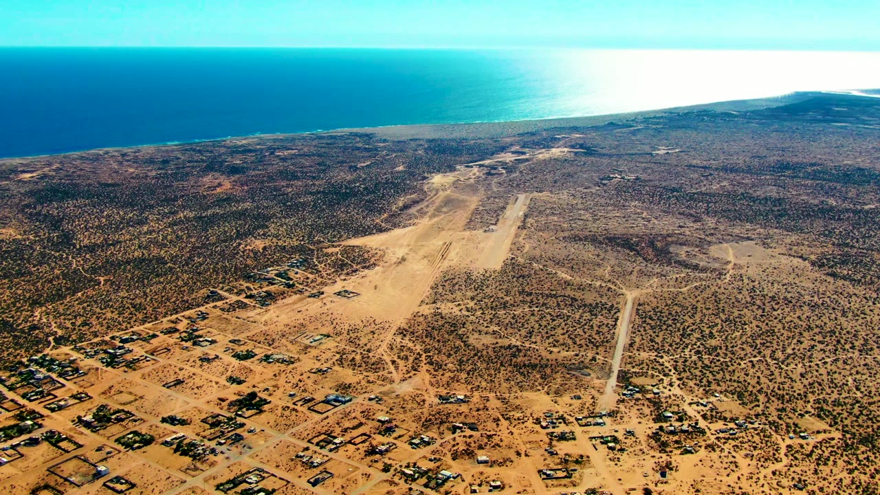 High aerial view over San Juanico, Baja California Sur, with a sandy runway centered in scrubby desert terrain, scattered buildings below, blue ocean beyond, and no visible people.
