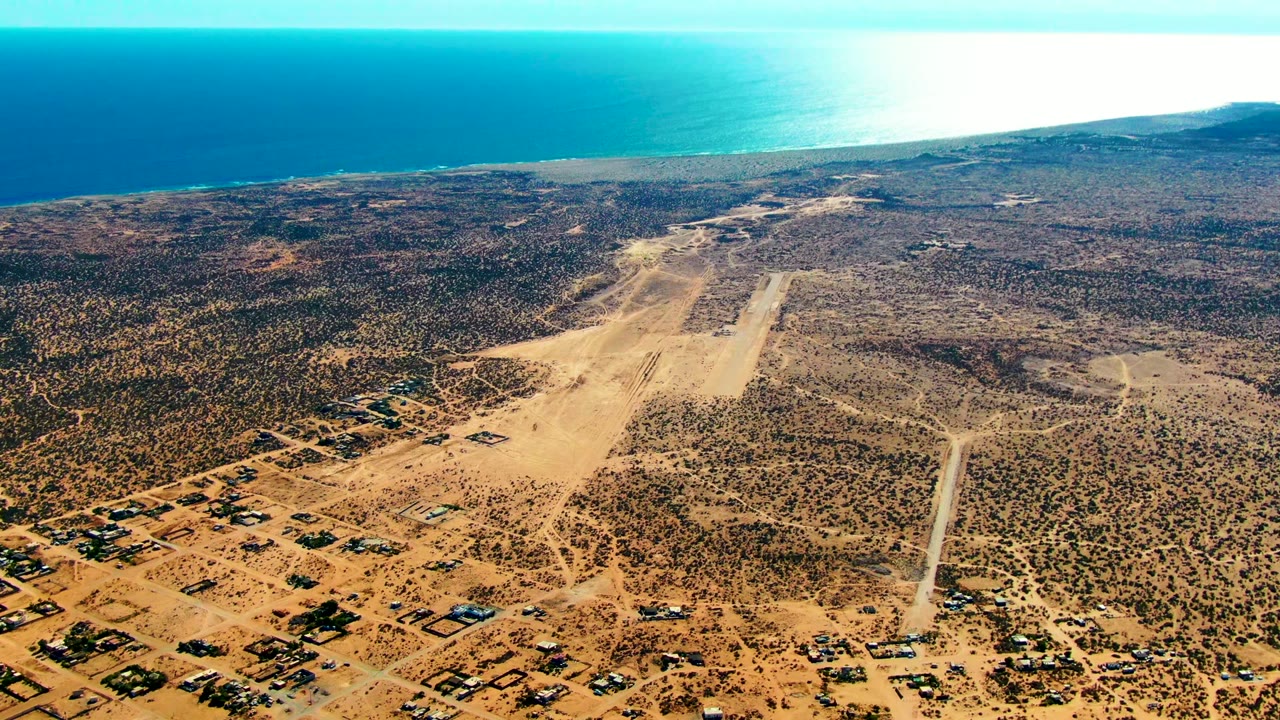 Wide drone view of the San Juanico coastal airfield with sandy runway, dry brown scrubland, a cluster of low structures near the lower edge, ocean in the background, and no visible people.