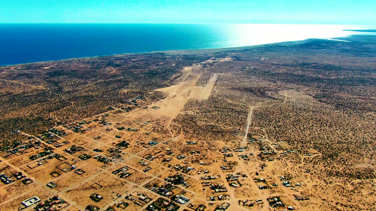 Aerial frame over San Juanico showing the runway slightly right of center, gridded sandy roads and homes below, scrubby desert around it, ocean in back, and no visible people.