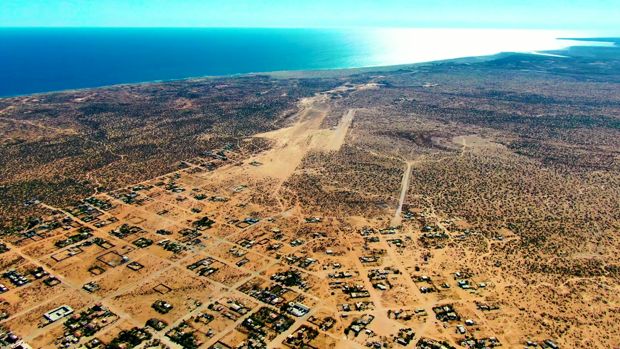 Static aerial over desert town blocks with runway and ocean horizon.