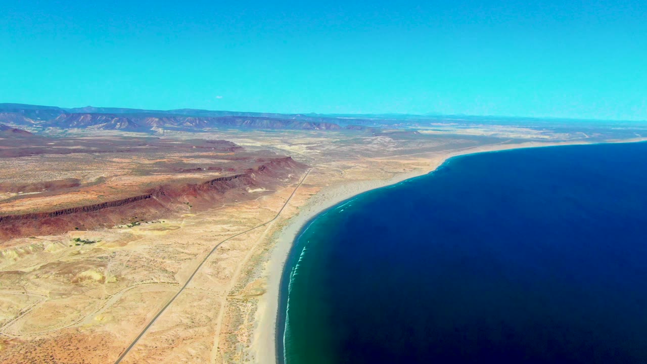 Opening aerial frame over San Juanico, Baja California Sur, with a curved sandy beach beside deep blue water, a desert road inland, distant mountains, and no visible people.