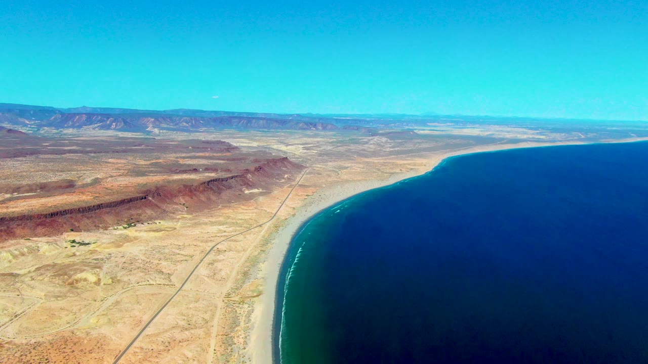 Wide aerial of arid coast with shoreline curve, inland road, ocean, and mountains.