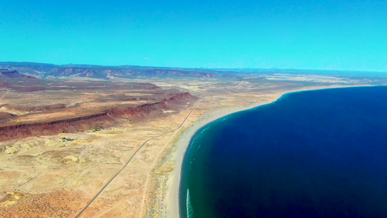 Aerial view over San Juanico, Baja California Sur, with a straight desert road, pale beach, deep blue ocean, distant mountains, and no visible people.
