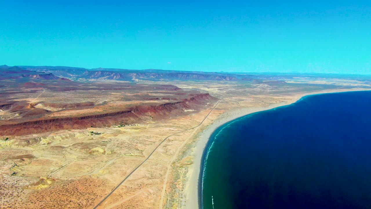 Aerial frame over San Juanico, Baja California Sur, showing tan desert, a coastal road, curved beach, deep blue ocean, distant mountains, clear sky, and no visible people.