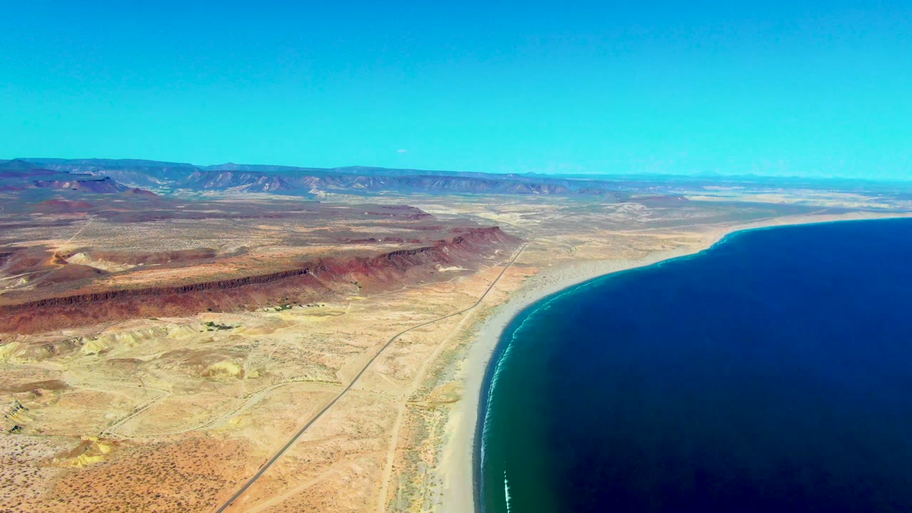 Aerial coastal desert with road parallel to the ocean and mountains beyond.