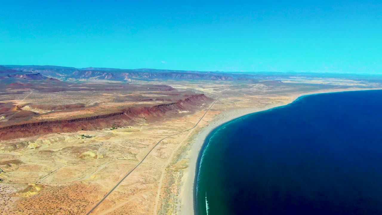 San Juanico aerial frame with dry ochre terrain, a narrow road near the beach, dark blue ocean to the right, low mountains in the distance, and no visible people.