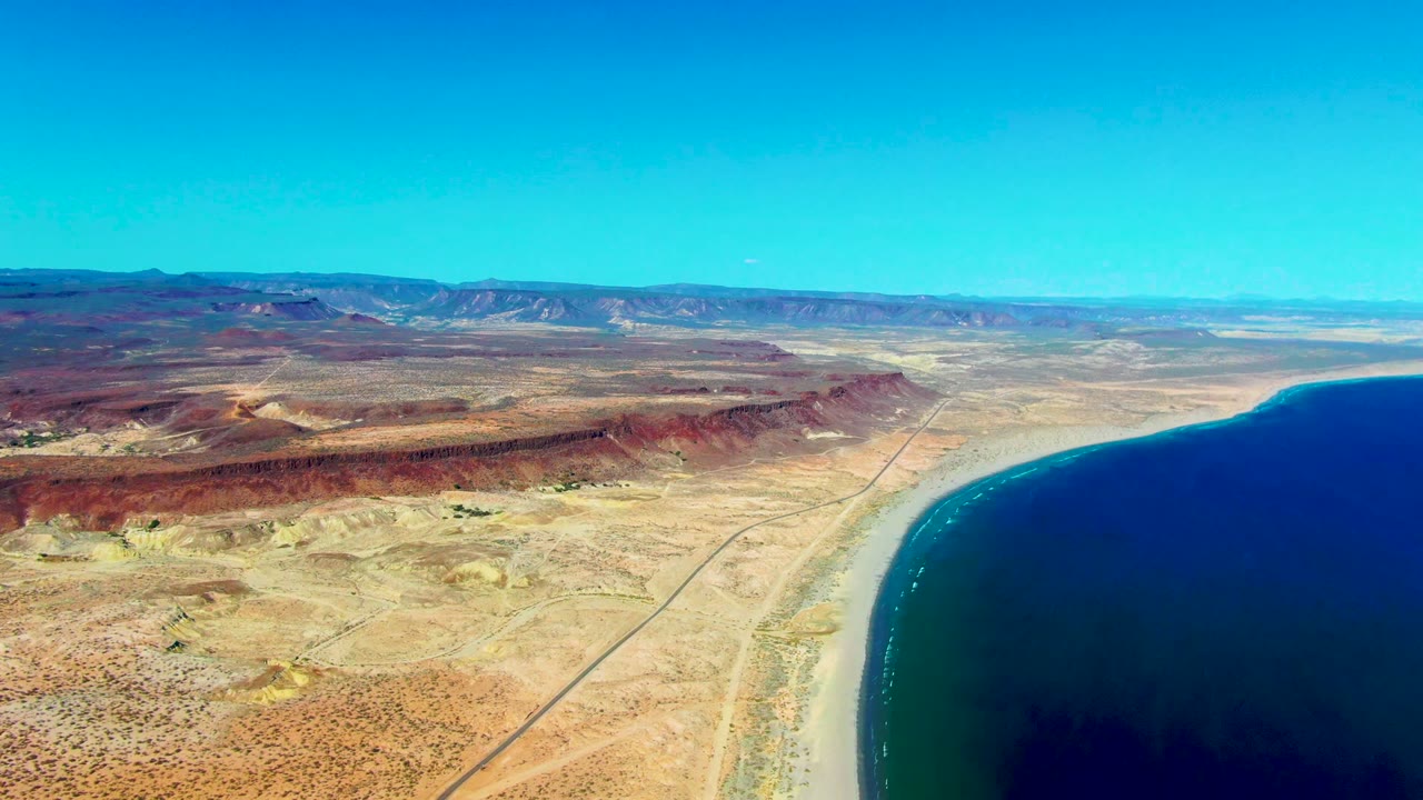 Opening frame of San Juanico, Baja California Sur showing blue ocean at right, pale shoreline, a straight road through arid desert, distant mountains, and no visible people.