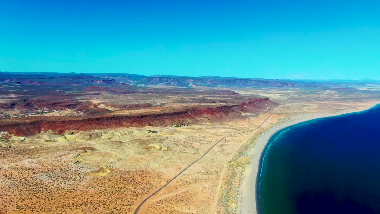 Aerial view over San Juanico, Baja California Sur, with a shoreline road crossing dry desert terrain beside dark blue ocean and no visible people.