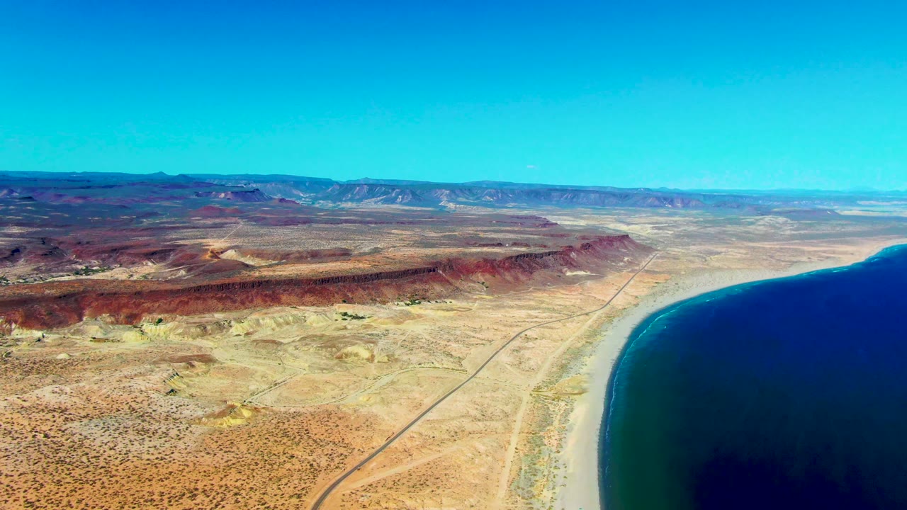 Aerial view in San Juanico with dark ocean filling the right side, a narrow beach curve, dry rocky ground, distant ridges, and no visible people.