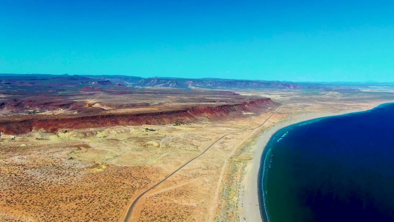 Opening aerial frame over San Juanico, Baja California Sur shows a desert road curving beside a sandy beach and dark blue ocean, with red ridges and no visible people.