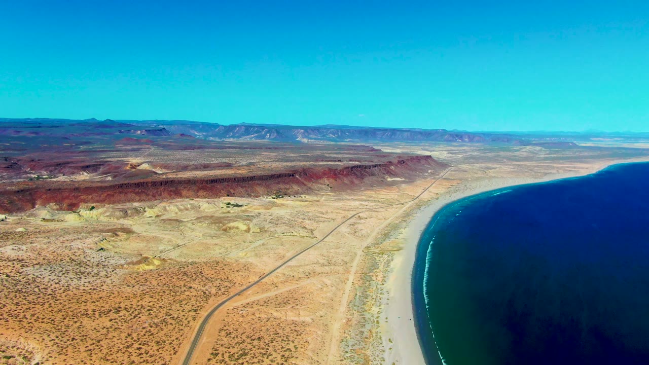 Opening aerial frame over San Juanico, Baja California Sur, with a desert road leading toward a pale beach, dark blue ocean at right, red ridges inland, and no visible people.