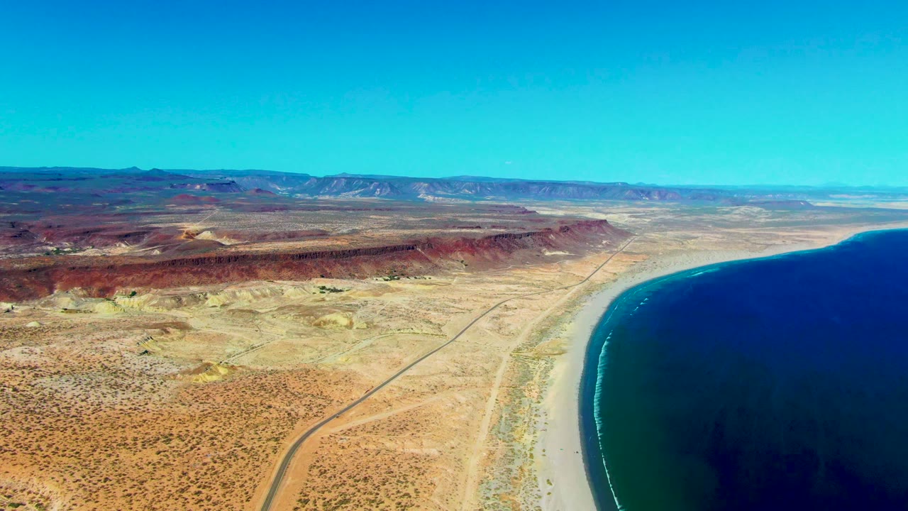 Second aerial frame over San Juanico, Baja California Sur, showing the road running through tan desert toward the shoreline, with ocean filling the right side and no visible people.