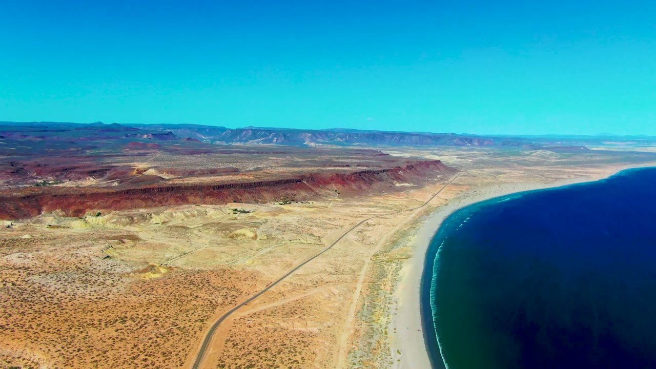 Aerial frame in San Juanico, Baja California Sur, with a stronger sweep of blue ocean on the right, pale shoreline below, dry desert road at left, and no visible people.