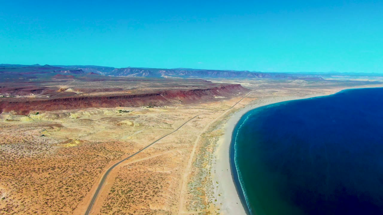 High aerial view in San Juanico, Baja California Sur, showing a desert road, pale beach, deep blue ocean, red-brown ridges, and no visible people at the start of the clip.