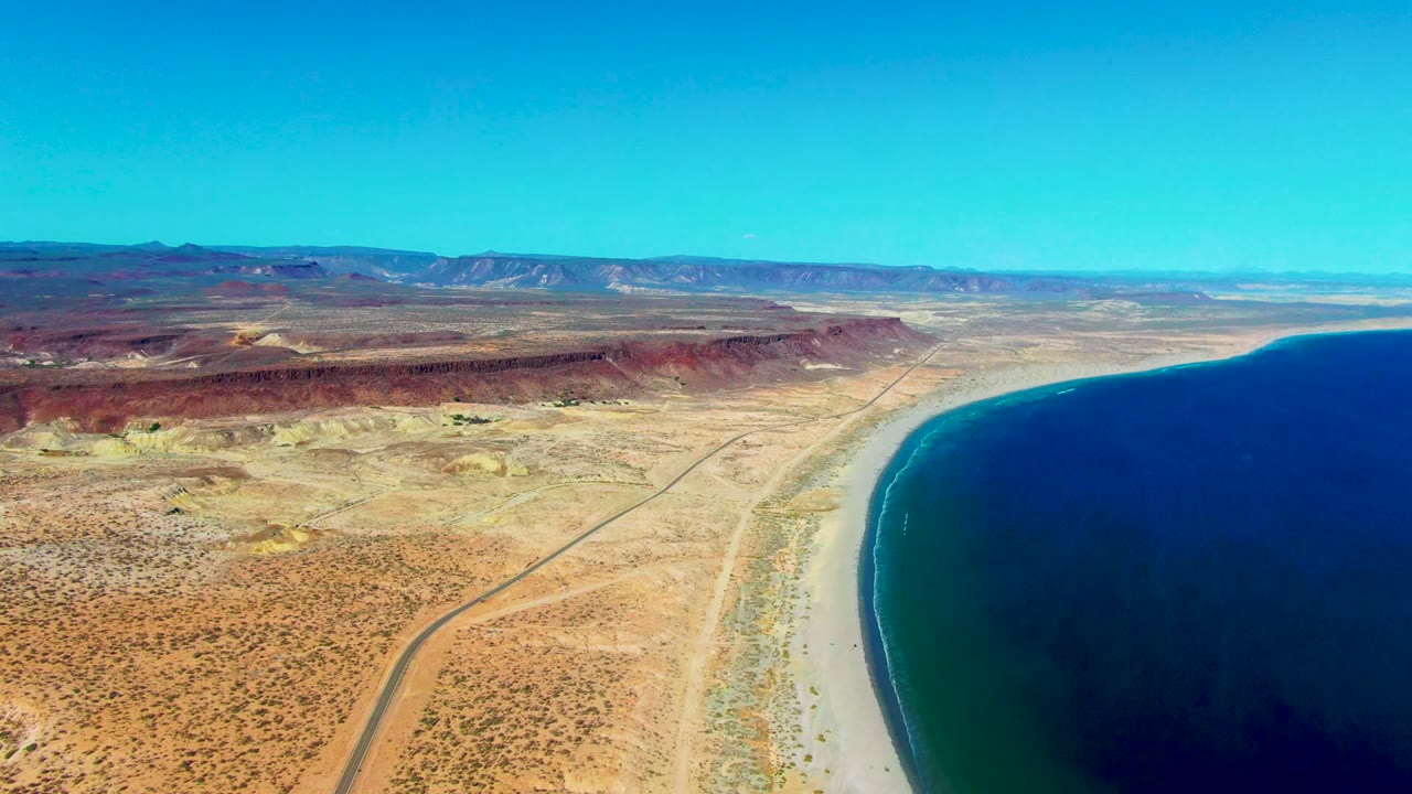 San Juanico aerial view showing more of the curving shoreline on the right, a straight desert road in the center, layered red ridges in the background, and no visible people.