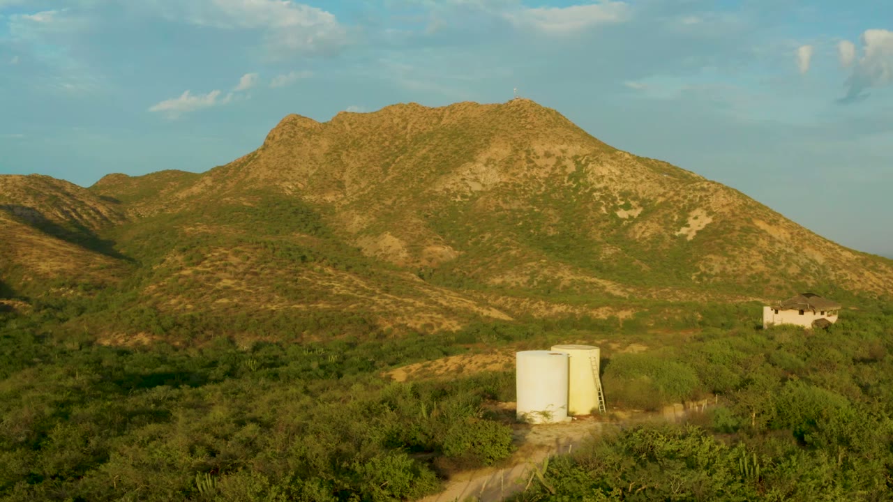 Wide view of a rugged green mountain slope in Cabo Pulmo, Baja California Sur, with a white cylindrical tank in the lower frame, a small pale building to the right, partly cloudy sky, and no visible people.
