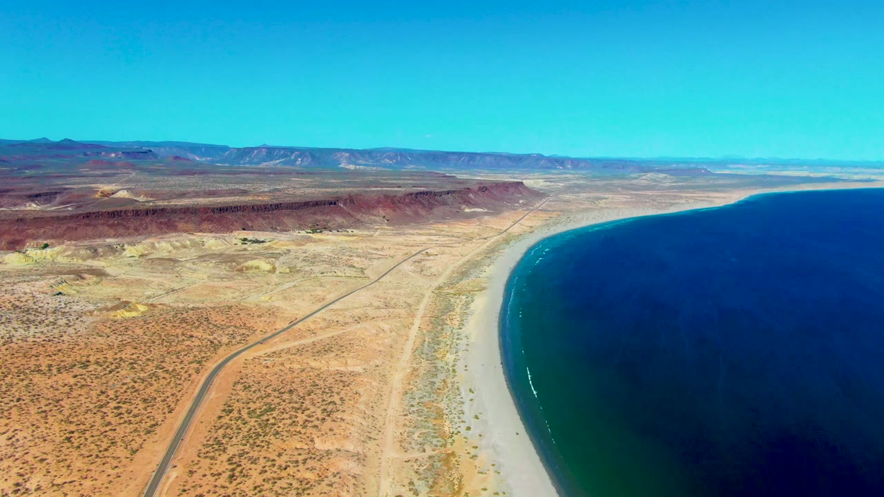 High aerial view over San Juanico, Baja California Sur, with a desert road crossing scrubland beside a curved sandy beach, deep blue ocean, distant ridges, and no visible people.