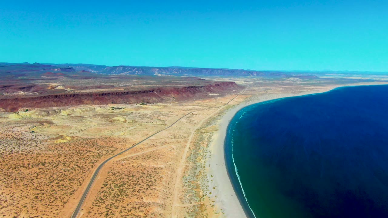 Wide aerial view over San Juanico, Baja California Sur, showing more of the inland desert road, red-brown ridge line, pale shoreline, dark blue ocean, and no visible people.