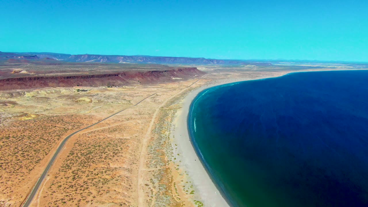 High aerial view over San Juanico, Baja California Sur, with a desert road at left, pale sand along the shore, deep blue water at right, distant mountains, and no visible people.