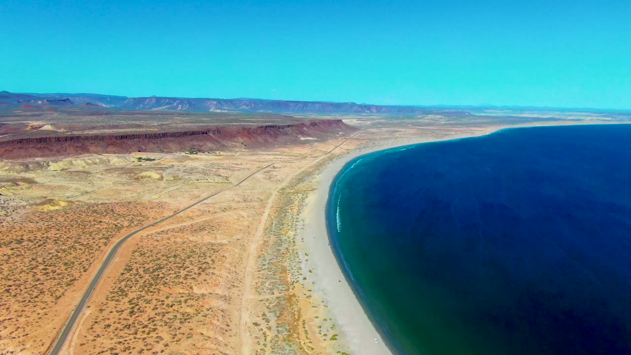 Desert coastline with road, beach, blue water, and mountain horizon