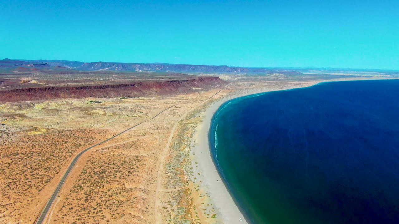 Wide aerial view of San Juanico coast with tan desert foreground, a thin road, a pale beach strip, saturated blue ocean, reddish inland ridges, and no visible people.