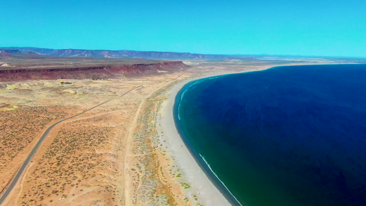 Coastal desert road curves near a long beach and dark blue water.