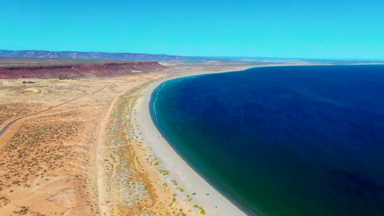 Aerial frame over San Juanico, Baja California Sur, showing a curving sandy shoreline, deep blue ocean, desert scrub, a parallel road, distant mountains, clear sky, and no visible people.