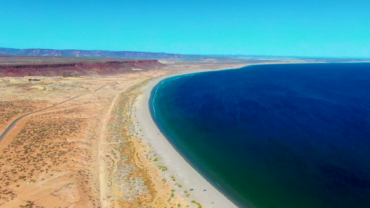 Elevated view of a pale beach curving along dark blue water and dry inland terrain.
