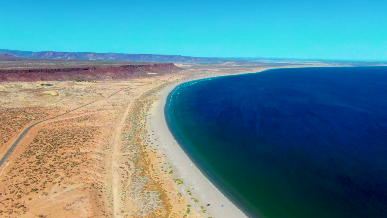 Desert road and sandy coast seen from above beside a broad expanse of blue water.
