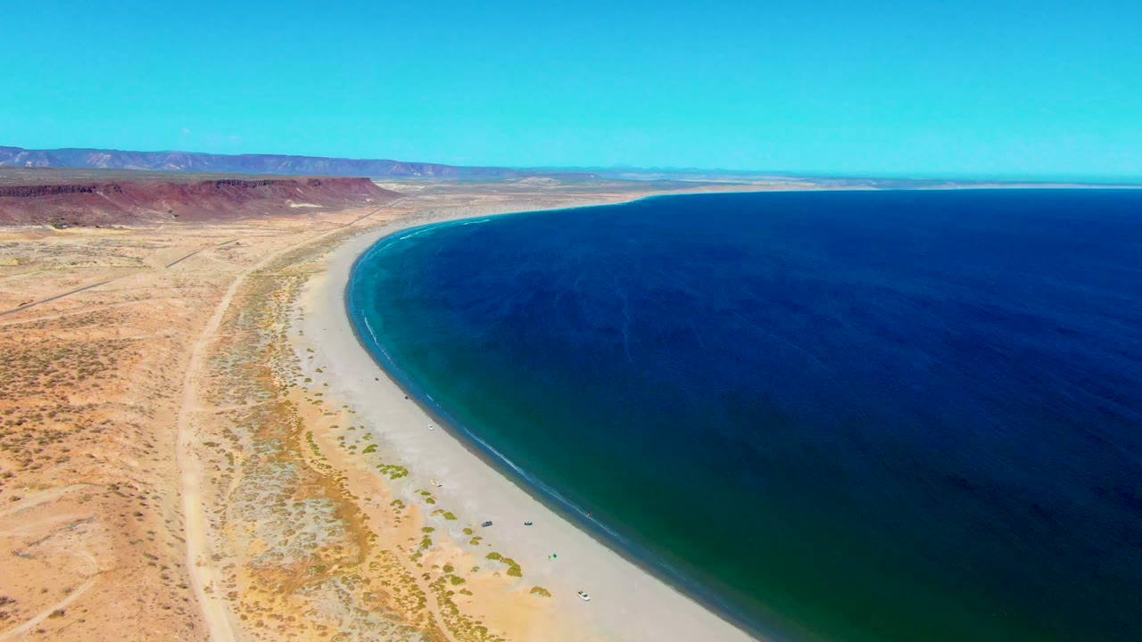 Aerial frame over San Juanico, Baja California Sur, showing a curved sandy shoreline, dry desert tracks, distant mesas, deep blue water, and no visible people.