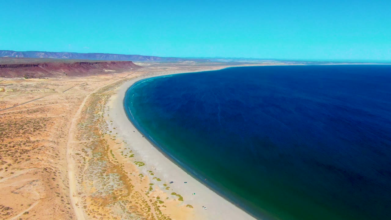 Curving desert shoreline with blue ocean on the right and pale beach below.