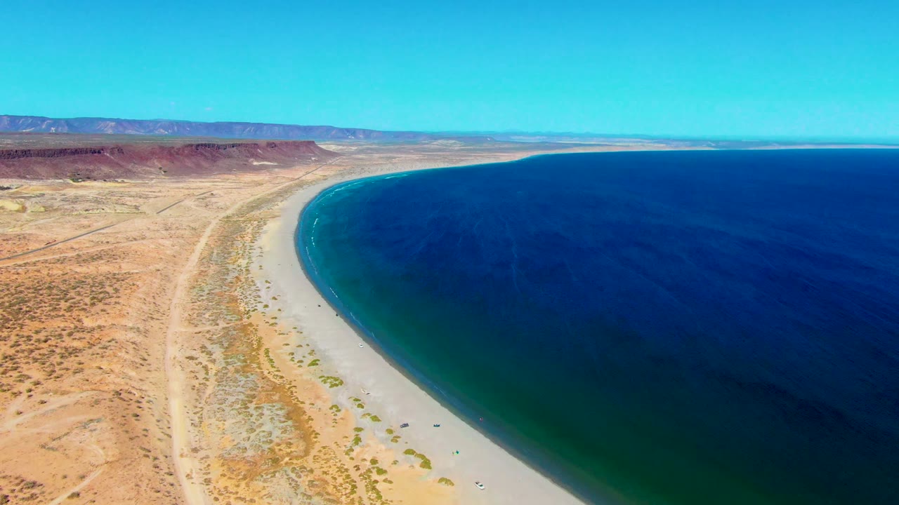 San Juanico aerial frame with a broad sweep of blue water, pale sand, sparse desert vegetation, long dirt tracks, distant red-brown mesas, and no visible people.