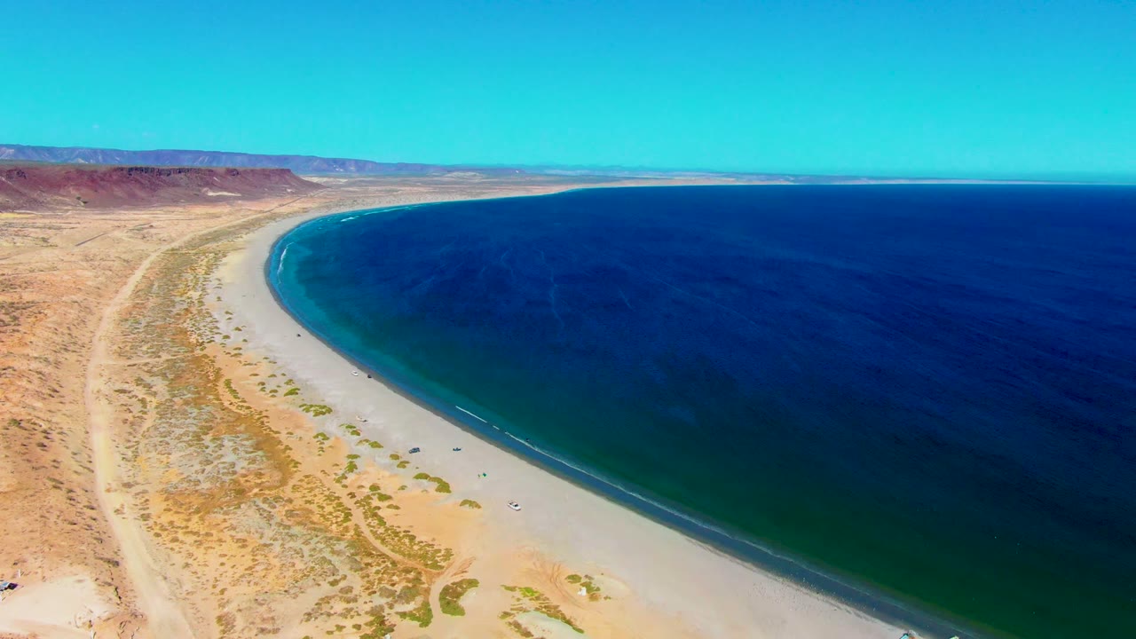 Aerial frame over San Juanico, Baja California Sur showing a pale sandy beach curving between dry scrubland and dark blue ocean, with tiny people visible on the shore.