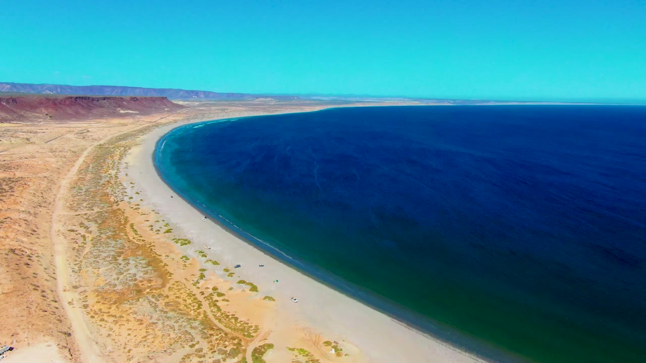 San Juanico beach appears from above with tan desert land on the left, a pale sand strip along the waterline, and broad blue ocean stretching to the horizon.