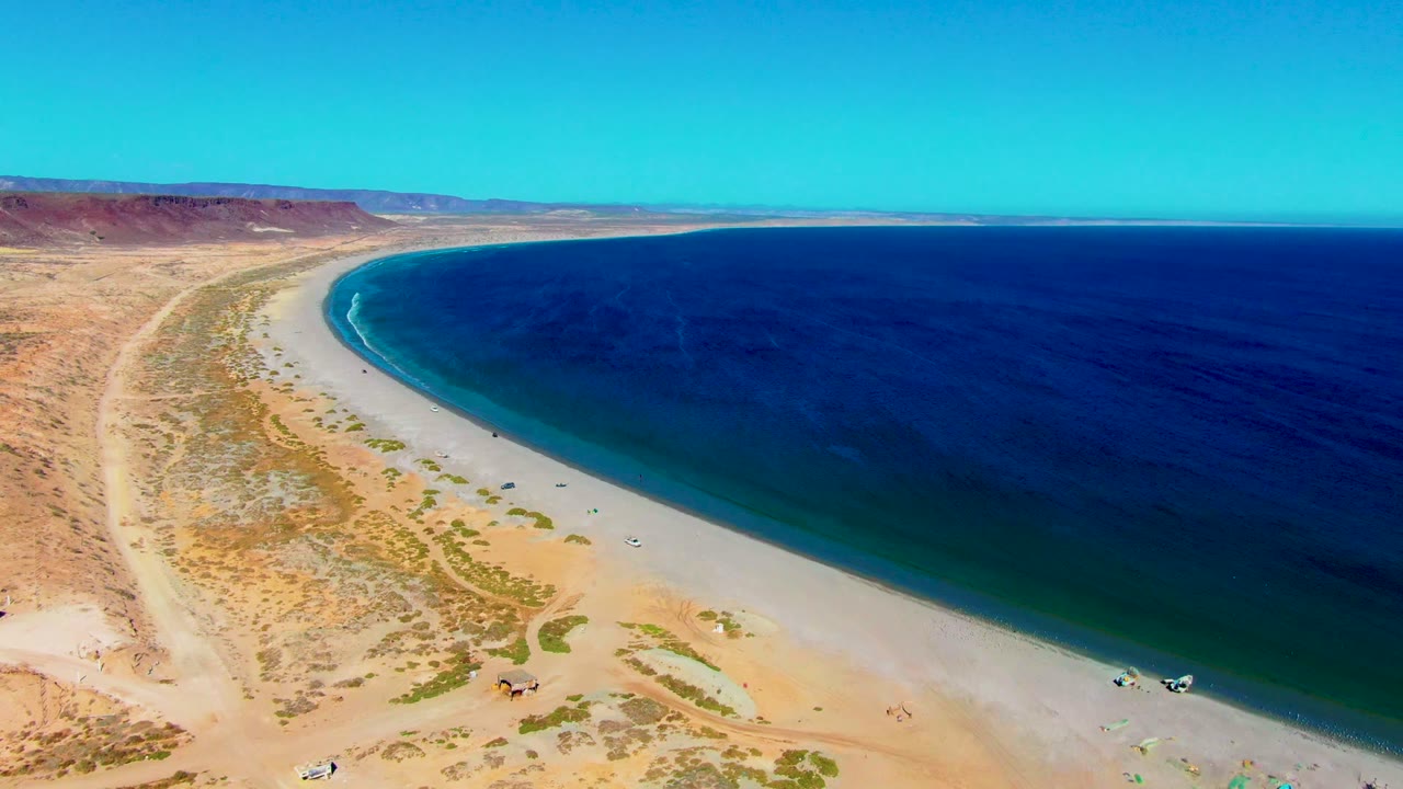 Opening aerial frame over San Juanico, Baja California Sur, with a pale curved beach, dark blue ocean, desert scrub, inland tracks, distant mesas, and no visible people.
