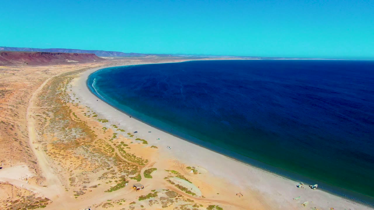 Wide drone view of shoreline, desert scrub, inland tracks, and blue ocean.