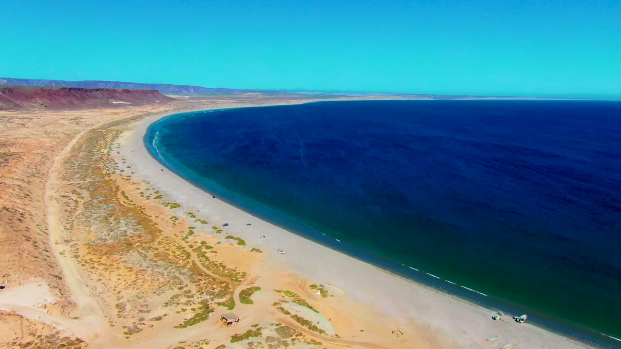 The two-second frame shows San Juanico's beach arc under clear light, with more shoreline visible to the right, deep blue water, dry desert flats, reddish mesa slopes, and no visible people.