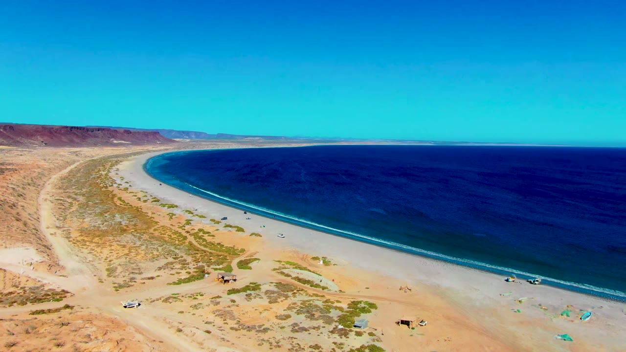 Aerial view over San Juanico, Baja California Sur, showing a curved sandy beach, blue ocean, desert scrub, dirt road, small vehicles, and tiny beachgoers near the lower shoreline.
