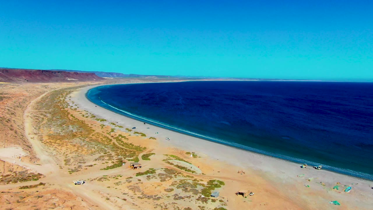 Wide drone view of sandy shoreline, blue water, desert road, and small beach activity.