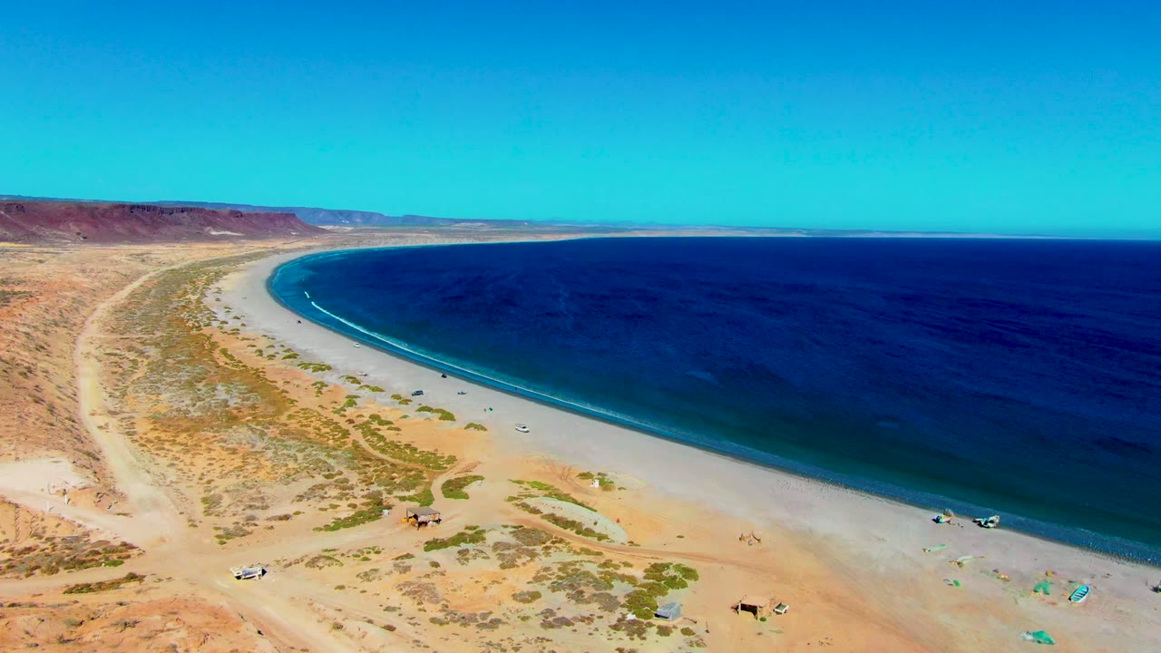 Aerial view of San Juanico beach with pale sand bending around the bay, dark blue water to the right, desert flats and ridges to the left, and tiny vehicles and beachgoers near the lower edge.